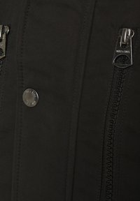 Gros plan sur un tissu noir avec un bouton-pression argenté et deux fermetures éclair noires avec des tirettes métalliques.