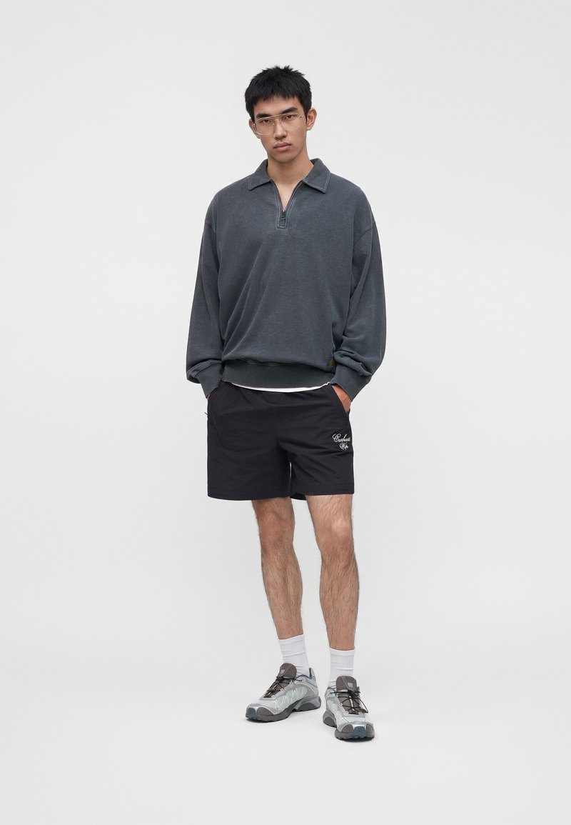 Young man wearing a dark grey zip-up sweatshirt, black shorts with white text, white socks, and grey sneakers, standing against a plain background.