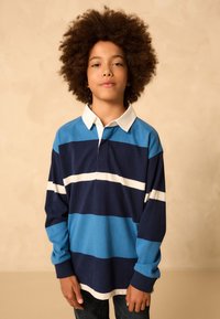 Ilgas rankovėmis polo marškinėliai mėlynos ir tamsiai mėlynos dryžuotos su baltais akcentais, klasikiniu apykakle ir laisvo silueto. Medvilninė audinys su raukšlėtais rankogaliais.