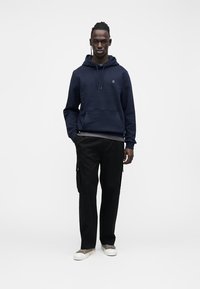 Tmavomodrá mikina s predným vreckom a sťahovacími šnúrkami, v kombinácii s čiernymi cargo nohavicami s bočnými vreckami a svetlými topánkami.