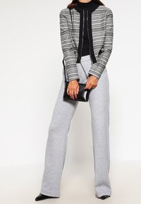 Veste cintrée texturée à motifs noir et blanc, col roulé noir, pantalons gris à jambes larges, et petit sac à main noir. Talons noirs pointus.