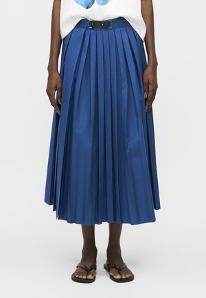 Άτομο με σκουρόχρωμη επιδερμίδα που φορεί μπλε πλισέ φούστα μήκους μέχρι τη μέση με μαύρη ζώνη, λευκό τοπ και μαύρα σανδάλια ανοιχτά στο δάχτυλο, στέκεται μπροστά σε απλό φόντο.