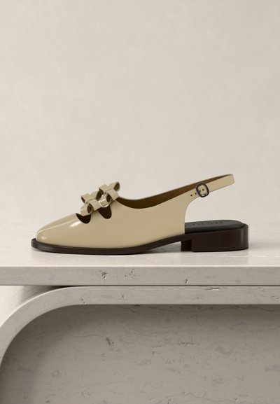 Beige flad slingback-sko med to dekorative sløjfer på toppen, firkantet tå og mørkebrun sål, vist på en lys marmorovertflade.