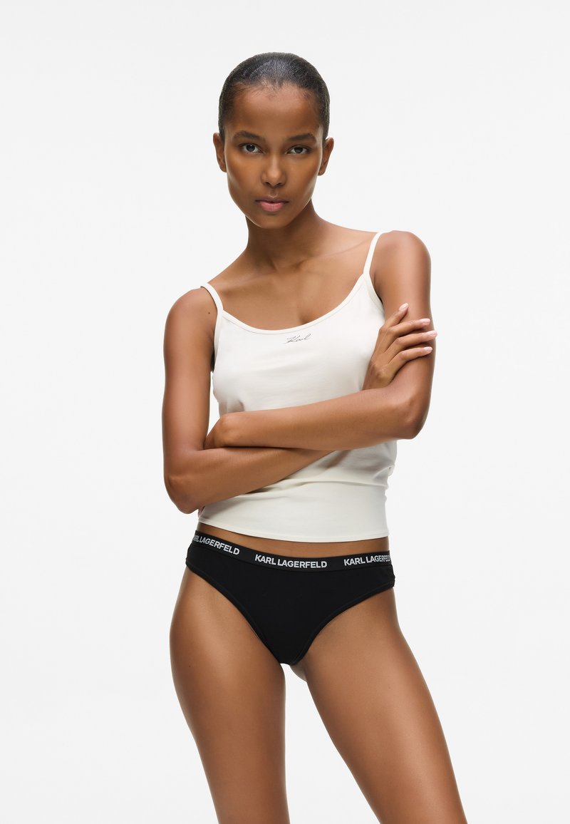 Kobieta z założonymi rękami, ubrana w białą koszulkę na ramiączkach i czarną bieliznę Karl Lagerfeld, na tle jednolitego białego tła.