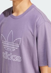 Person som bär en lila Adidas t-shirt med en stor vit konturlogo på bröstet, visad från halsen till mitten av överkroppen.