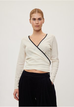 Femme aux cheveux blonds avec une raie au milieu portant un haut crème à manches longues style cache-cœur avec des bordures noires et un pantalon noir, debout devant un fond uni.