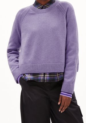 Person iført en lavendelfarvet striktrøje over en lilla ternet skjorte, med sorte bukser og den ene hånd i lommen mod en ensfarvet baggrund.