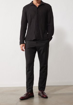 Chemise texturée noire à manches longues avec un col, associée à un pantalon noir et des chaussures en cuir bordeaux foncé.