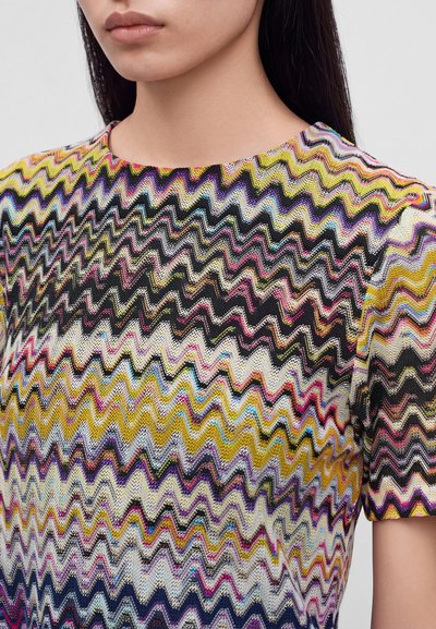 Femme portant un haut à manches courtes avec un motif en zigzag coloré dans des tons jaunes, noirs, roses et bleus.
