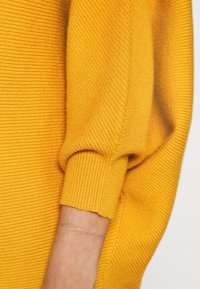 Gros plan sur le bras d'une personne portant un pull côtelé jaune avec des poignets ajustés et un fin tatouage léger visible au poignet.