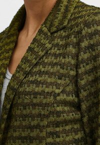Grüner strukturierter Blazer mit kariertem Muster, ausgestattet mit einem Revers und einer formvollen Silhouette aus einer Mischung gewebter Materialien.