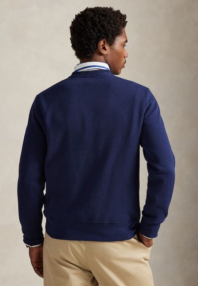 Mann iført marineblå genser over stripete skjortekrage og beige bukser, stående med den ene hånden i lomma, med ryggen mot en nøytral bakgrunn.