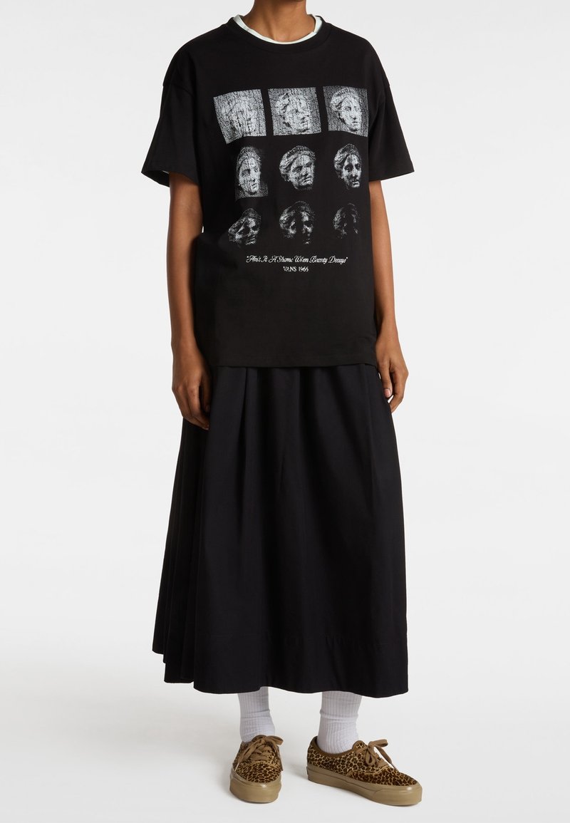 Personne portant un t-shirt noir à motifs avec des visages abîmés, une jupe midi noire, des chaussettes blanches et des baskets à imprimé léopard sur fond blanc.