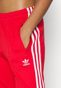 Röda Adidas sweatpants med vita sidostickningar, en logotyp på vänstra låret och fickor. Materialet ser slätt och sportigt ut.