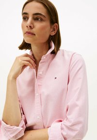 Camisa de algodón a rayas en rosa claro y blanco con cuello de botón y un pequeño logo bordado en el lado izquierdo del pecho. Mangas largas arremangadas.