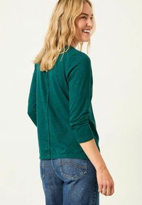 Teal green long-sleeve top with a back seam detail, worn with blue denim jeans. Soft fabric with a relaxed fit and round neckline.