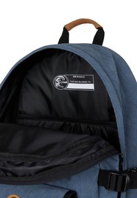 Blauwe corduroy rugzak met een zwart interieur, inclusief een label voor personalisatie en een ritscompartiment. Bevat een bruine handgreep.