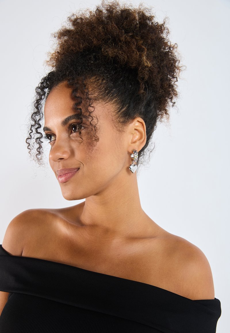Pendientes de plata con un diseño de corazón y acentos decorativos. El modelo lleva una blusa negra sin hombros. El fondo es blanco liso.
