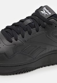 Black leather sneaker featuring textured upper, thick laces, perforations for breathability, and rubber sole with patterned tread. Reebok logo on tongue.