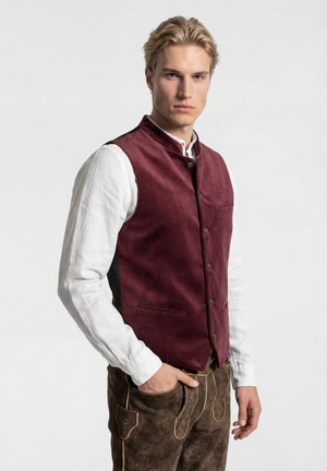 Junger Mann mit blondem Haar, der ein weißes Hemd, eine burgunderfarbene knöpfbare Weste und braune traditionelle Lederhosen trägt, mit der Hand in der Tasche.