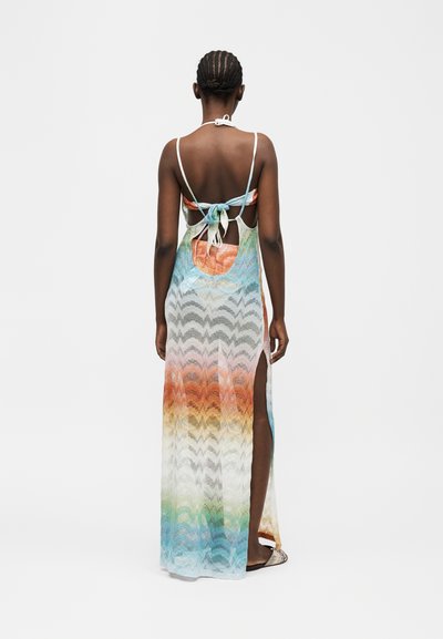 Γυναίκα με πλεξουδάκια στα μαλλιά, φορώντας πολύχρωμο, αμάνικο καλοκαιρινό φόρεμα με ανοίγματα στην πλάτη και σχίσιμο στο πλάι, στέκεται μπροστά σε ένα απλό λευκό φόντο.