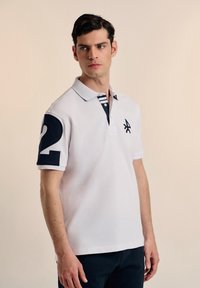 Camisa polo blanca con cuello azul marino y detalles a rayas. Presenta un gran número "2" en azul marino en la manga izquierda y un emblema en el pecho.