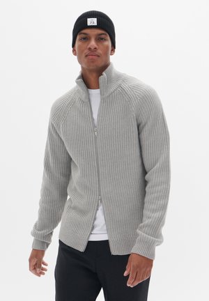 Szary prążkowany kardigan zapinany na zamek z wysokim kołnierzem i długimi rękawami, w zestawieniu z białą koszulą. Model nosi czarną czapkę beanie.