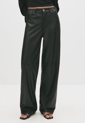 Pantalones anchos de cuero sintético negro combinados con sandalias negras con tiras, top corto no completamente visible, de pie frente a un fondo liso.