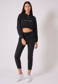 Schwarze, cropped Kapuzenjacke mit Reißverschluss und Kordelzug, kombiniert mit schwarzen Jogginghosen mit elastischen Bündchen und kontrastierenden Seitenstreifen. Weiße Sneaker.