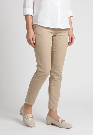 Person trägt beiger, schmal geschnittener Hose, weißes Hemd mit Knöpfen und hellbeige Loafers mit goldener Kettendetailierung, steht vor weißem Hintergrund.