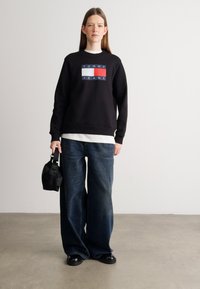 Mustat huppari, jossa on "Tommy Jeans" -logo suorakulmaisen värilohkon päällä. Yhdistetty rentoihin sinisiin farkkuihin ja pieneen mustaan käsilaukkuun.