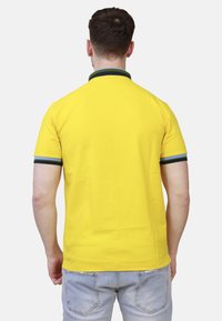 Maglietta polo gialla con colletto verde e polsini neri, caratterizzata da una texture liscia e maniche corte.