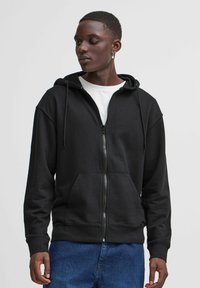 Black zip-up hoodie in cotton blend, featuring a drawstring hood, front pockets, and ribbed cuffs. Worn over a white t-shirt, casual fit.