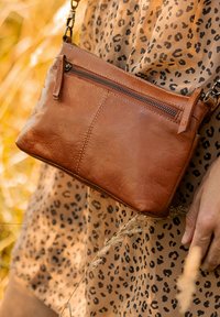 Borsa a tracolla in pelle marrone con tasca con zip, texture liscia e forma rettangolare, indossata su un vestito beige con macchie di leopardo.