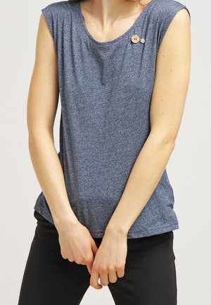 Haut sans manches bleu chiné avec deux petits boutons en bois près de l'encolure, porté avec un pantalon noir, mains jointes devant.