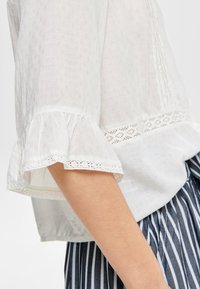 Blouse blanche en tissu texturé, avec des manches à volants et des détails en dentelle à l'ourlet, associée à un tissu rayé en dessous.