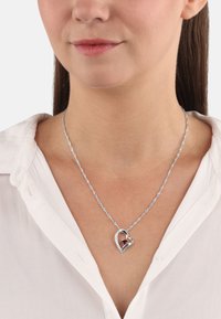 Collar de cadena trenzada en tono plateado que presenta un colgante en forma de corazón con una piedra preciosa transparente y un orbe rojo en el centro.