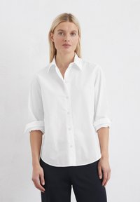 Camicia bianca con bottoni realizzata in cotone. Presenta un colletto, maniche arrotolate e orlo dritto, abbinata a pantaloni scuri. Texture liscia.