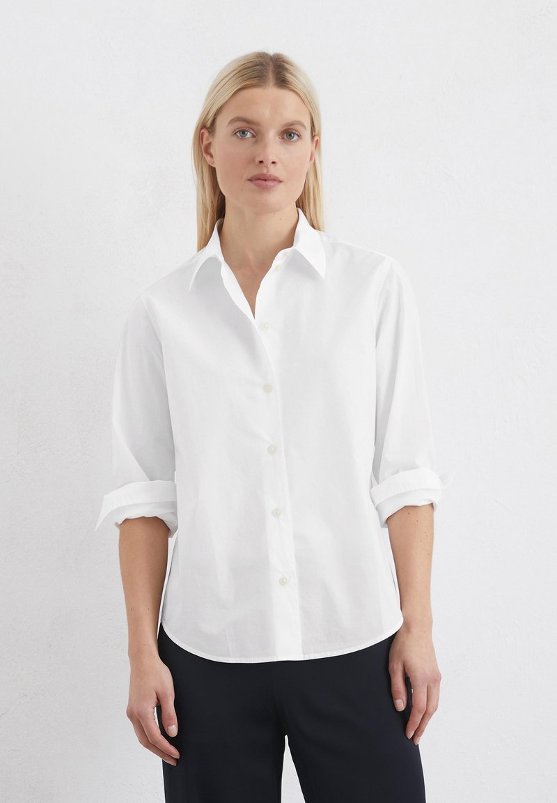 Camicia bianca con bottoni realizzata in cotone. Presenta un colletto, maniche arrotolate e orlo dritto, abbinata a pantaloni scuri. Texture liscia.