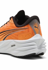 Zapatilla deportiva naranja con un upper de malla texturizada, acentos negros y una suela blanca acolchada. Presenta un logo plateado y elementos de diseño estilizados.