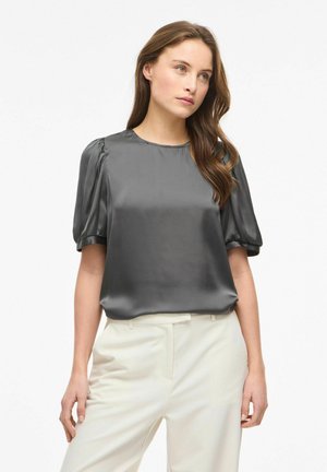 Hosszú barna hajú nő fényes szürke pöttyös ujjú blúzt és magas derekú fehér nadrágot visel, fehér háttér előtt áll.