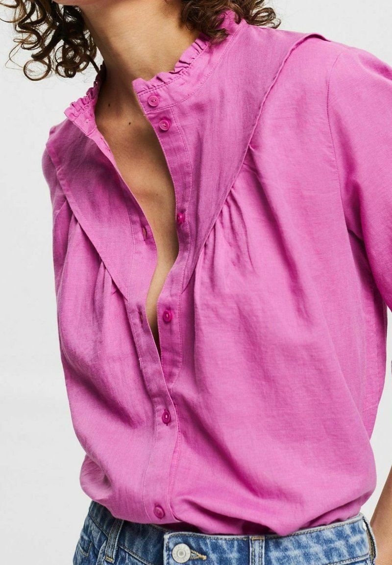 Chemise rose à manches longues avec un col froncé et de légères fronces au niveau de la poitrine ; dispose de boutons de couleur assortie et d'une texture de tissu douce.