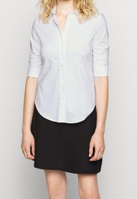 Camicia bianca con bottone e maniche lunghe arrotolate fino a tre quarti, abbinata a una gonna nera con cuciture verticali. Texture liscia.