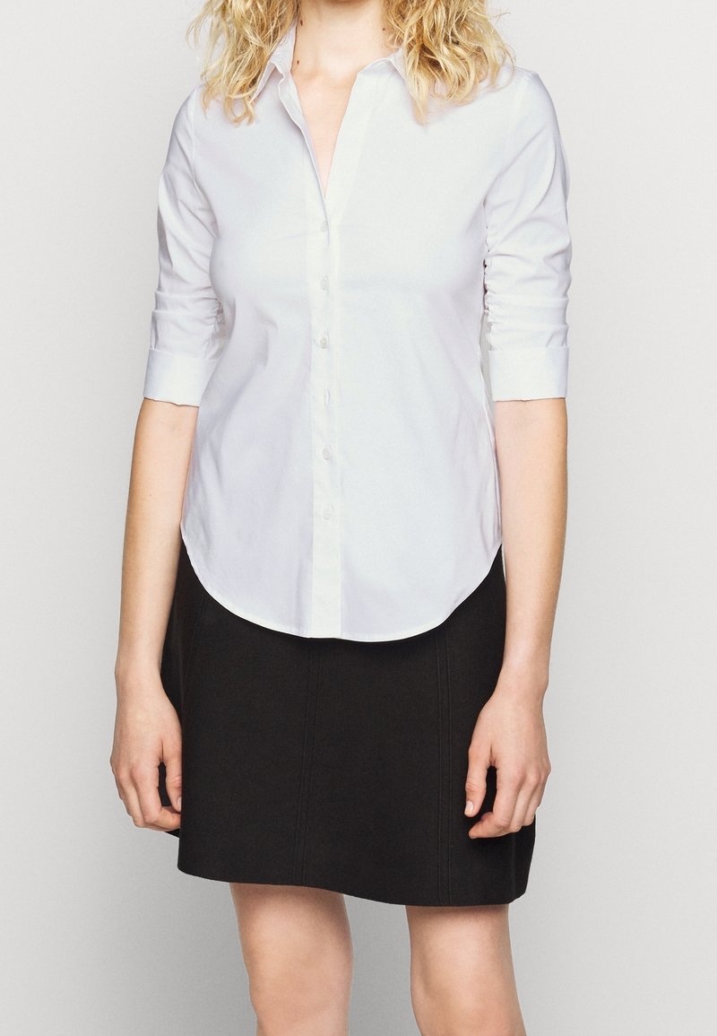 Camicia bianca con bottone e maniche lunghe arrotolate fino a tre quarti, abbinata a una gonna nera con cuciture verticali. Texture liscia.
