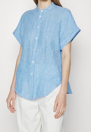 Woman wearing light blue short-sleeve button-up shirt with white vertical stripes and white pants, standing against a white background.