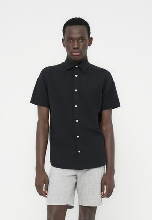 Crna košulja s kratkim rukavima od pamučne mješavine, s ovratnikom i bijelim gumbima; uparena sa svijetlosivim hlačicama.