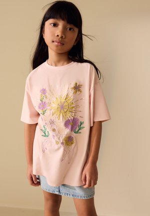 Mädchen mit langen schwarzen Haaren trägt ein hellrosa übergroßes T-Shirt mit gestickter Sonne, Blumen und Pferd, kombiniert mit einem hellblauen Jeansrock.
