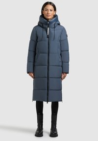 Fodrad lång kappa i mörkblått, med dragkedja, justerbar huva och stora fickor. Tillverkad av ett slätt, glänsande tyg.