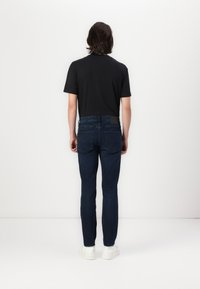 Jeans blue din denim pentru bărbați, cu croi slim, dotate cu o etichetă din piele la spate, stilizate cu un tricou negru și adidași albi.