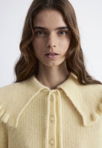 Helles gelbes Strickoberteil mit großem Kragen, Knopfverschluss und strukturiertem Stoff. Verfügt über gefranste Akzente an den Schultern.